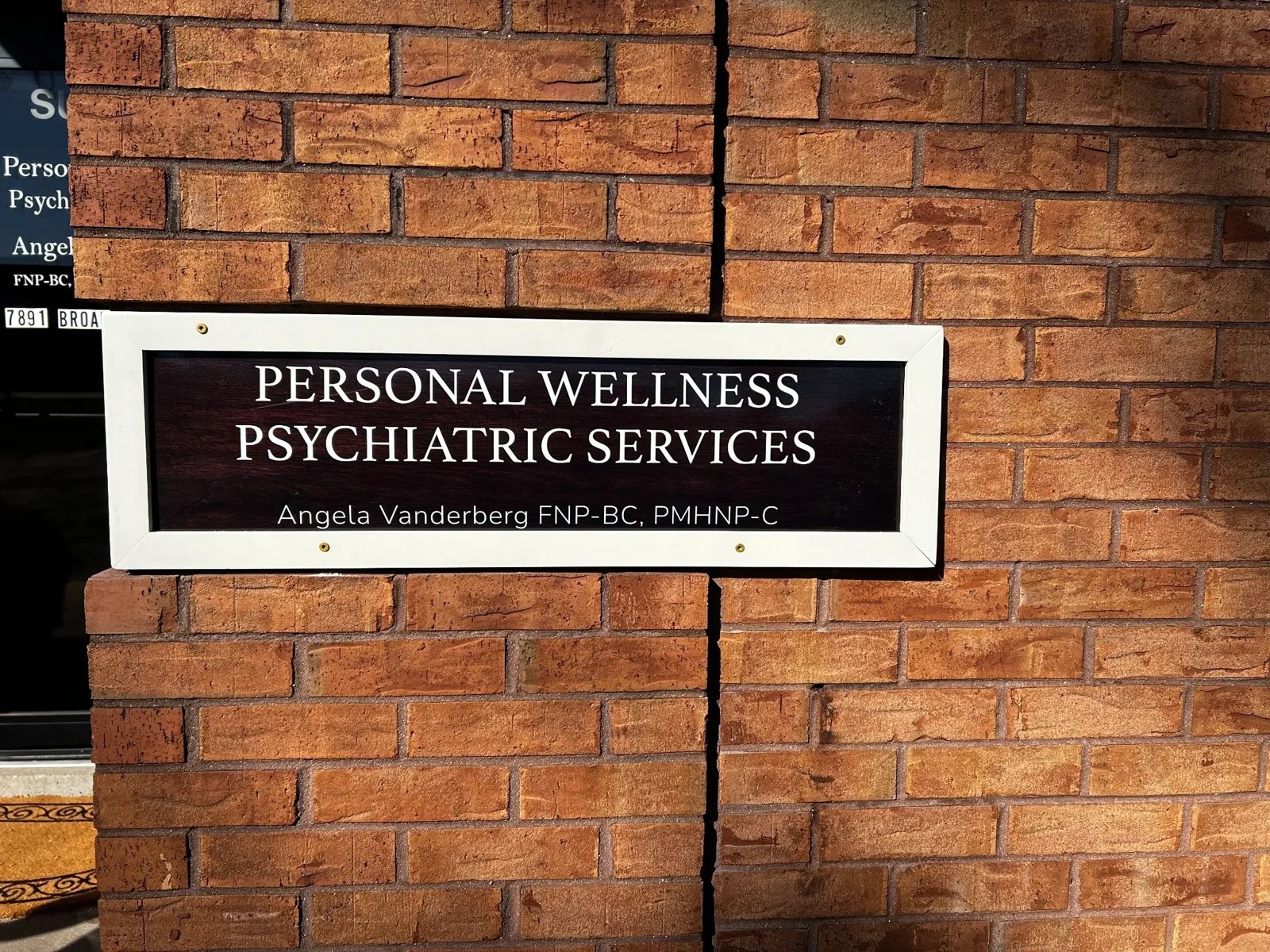 The white-framed Personal Wellness Psychiatric Services sign on the brick wall, with Angela Vanderberg FNP-BC, PMHNP-C below the name